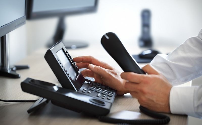 Person hält Telefonhörer und tippt Nummer auf einem schwarzen Bürotelefon am Schreibtisch